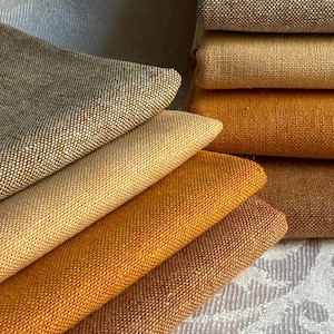 Op de afbeelding: Een stapel gevouwen stofstalen in verschillende tinten bruin, beige en goudgeel. Het materiaal heeft een zichtbare geweven textuur met een consistent rasterpatroon op elk stuk, wat een verloop van warme aardetinten laat zien.