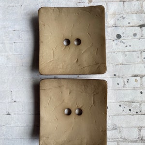 May include: Two brown square buttons with a textured surface and two holes in the center.