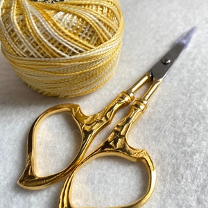 May include: A spool of yellow and white embroidery floss with the text "Double Gassed & Mercerized" and "8m/8yd" on the label. A pair of gold heart-shaped embroidery scissors.