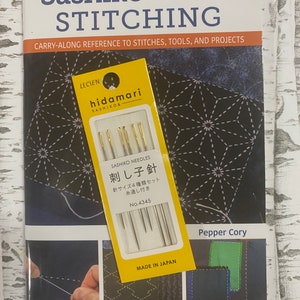 May include: A book titled "Pocket Guide to Sashiko and Boro Stitching" with a yellow package of "Sashiko Needles" on top. The book cover features a dark blue geometric pattern and the text "Carry-Along Reference to Stitches, Tools, and Projects". The needle package is labeled "Made in Japan".
