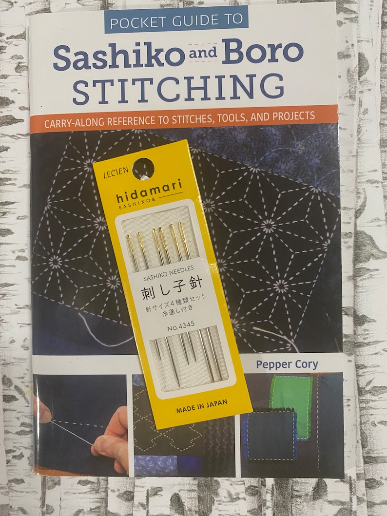 May include: A book titled "Pocket Guide to Sashiko and Boro Stitching" with a yellow package of "Sashiko Needles" on top. The book cover features a dark blue geometric pattern and the text "Carry-Along Reference to Stitches, Tools, and Projects". The needle package is labeled "Made in Japan".