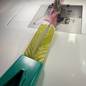 May include: A teal plastic bias tape maker tool feeding a strip of fabric into a sewing machine. The fabric features a light green leaf pattern on one side and a solid pink color on the other, with a white batting layer visible in the center.