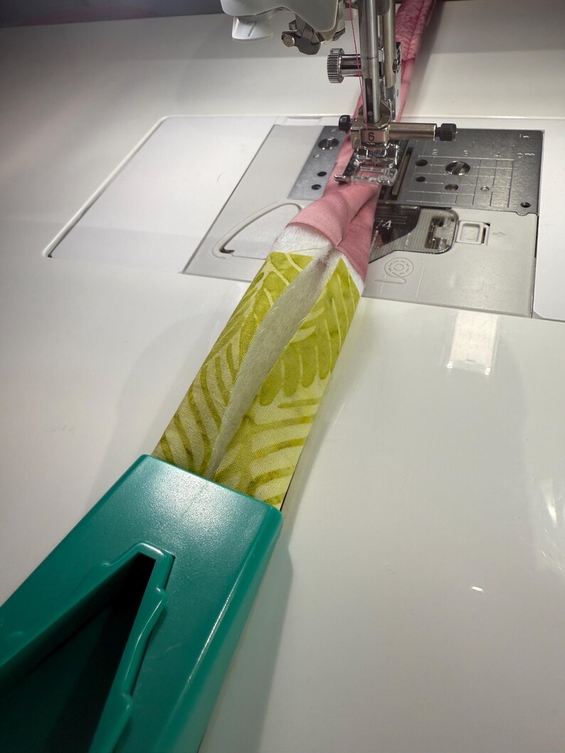 May include: A teal plastic bias tape maker tool feeding a strip of fabric into a sewing machine. The fabric features a light green leaf pattern on one side and a solid pink color on the other, with a white batting layer visible in the center.