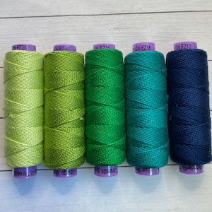 Puede incluir: Cinco carretes de hilo de bordar en tonos de verde y azul. Los carretes est&aacute;n etiquetados con el nombre de la marca "WonderFil" y el c&oacute;digo de color "Col. EZ30", "Col. EZ34", "Col. EZ36", "Col. EZ12" y "Col. EZ36".