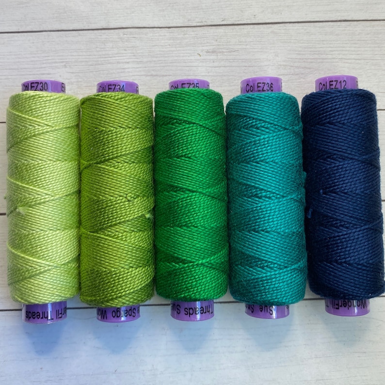 Puede incluir: Cinco carretes de hilo de bordar en tonos de verde y azul. Los carretes est&aacute;n etiquetados con el nombre de la marca "WonderFil" y el c&oacute;digo de color "Col. EZ30", "Col. EZ34", "Col. EZ36", "Col. EZ12" y "Col. EZ36".