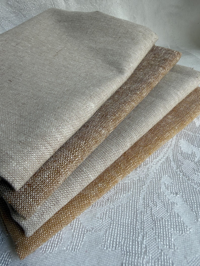 K&ouml;nnte beinhalten: Ein Stapel aus vier gefalteten Stoffmustern mit einer sichtbaren Webstruktur. Die Farben reichen von hellem Beige und Haferfarben bis hin zu Mittelbraun und einem tieferen Goldbraun, was eine nat&uuml;rliche, erdige Farbpalette unterstreicht.
