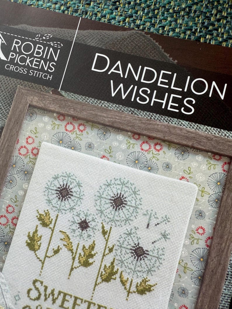 May include: Cross-stitch art titled "Dandelion Wishes" by Robin Pickens. The framed artwork features a light blue background with embroidered dandelions in shades of blue, brown, and green. The frame is a light brown color.
