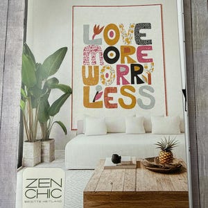 May include: A magazine page featuring a living room scene. A large wall hanging with the words "LOVE MORE WORRY LESS" in colorful, patterned letters hangs above a white sofa. A wooden coffee table holds a pineapple and decorative items.