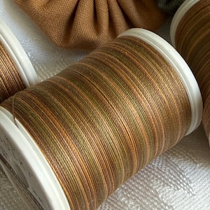 May include: A spool of variegated sewing thread featuring a blend of brown, tan, and olive green tones wound onto a white plastic cylinder. The thread displays a multi-colored striped appearance, and the spool base shows partial text reading SULKY.