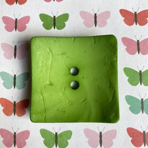 May include: A square green button with two holes. The button has a textured surface and a slightly faded look.