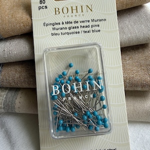 K&ouml;nnte beinhalten: Eine Packung mit 80 Bohin France Murano Glaskopfstecknadeln in T&uuml;rkisblau. Die Stecknadeln bestehen aus vernickeltem Stahl und messen 30 mm mal 0,60 mm. Die Verpackung ist wei&szlig; mit goldenem Text und einem durchsichtigen Kunststoffgeh&auml;use.