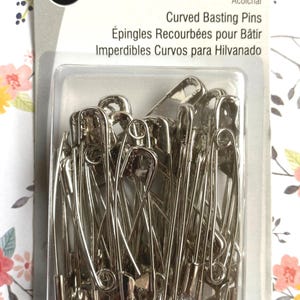 May include: A package of Dritz brand curved basting pins for quilting. The silver-colored metal pins are contained within a clear plastic blister pack attached to a cardboard backing that displays the product name in English, French, and Spanish.