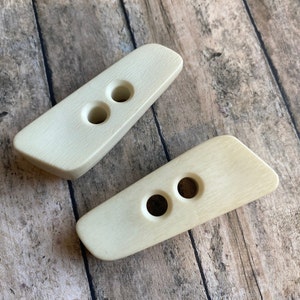 May include: Two off-white, wooden, rectangular buttons with two holes each. The buttons are on a wooden surface.