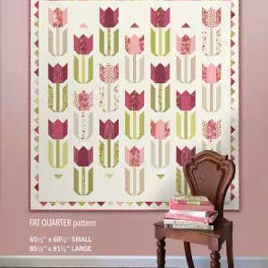 May include: A quilt with a tulip design in shades of pink, green, and cream hangs on a wall. The quilt is labeled "TULIP SEASON" and is available in small (65.5" x 68") and large (85.5" x 91.5") sizes. A wooden chair with books sits in front of the quilt.