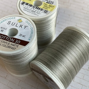 May include: Three spools of Sulky Cotton 30 thread in a neutral, light gray color. The spools are labeled with details including the thread weight, needle size, and yardage. The thread is designed for embroidery, quilting, and overlock projects.