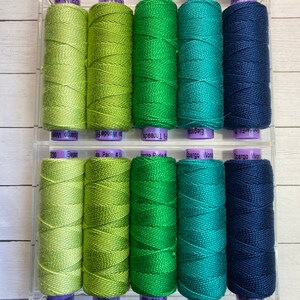 Puede incluir: Una colecci&oacute;n de 10 carretes de hilo de bordar en tonos verdes y azules. Los carretes est&aacute;n dispuestos en dos filas de cinco. Los carretes est&aacute;n etiquetados con los nombres de marca "WonderFil", "Fl Threads", "Elegance", "Perle #5", "Spargo Won" y "Spargo Wor".