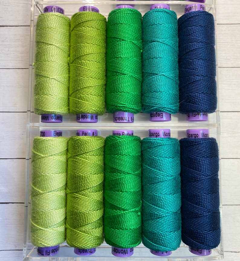 Puede incluir: Una colecci&oacute;n de 10 carretes de hilo de bordar en tonos verdes y azules. Los carretes est&aacute;n dispuestos en dos filas de cinco. Los carretes est&aacute;n etiquetados con los nombres de marca "WonderFil", "Fl Threads", "Elegance", "Perle #5", "Spargo Won" y "Spargo Wor".