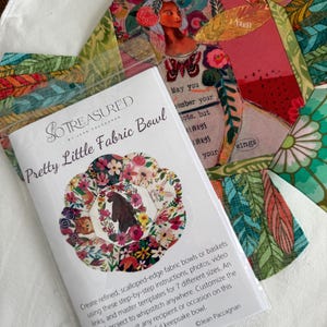 May include: A package for a "Pretty Little Fabric Bowl" by So Treasured, featuring a floral design. The package is on a white surface, with colorful fabric pieces and a decorative artwork with a woman and text.