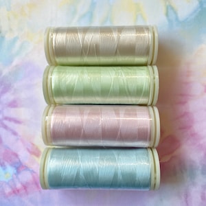 Op de afbeelding: Vier spoelen garen in pastelkleuren: beige, lichtgroen, roze en lichtblauw. De spoelen zijn op elkaar gestapeld.