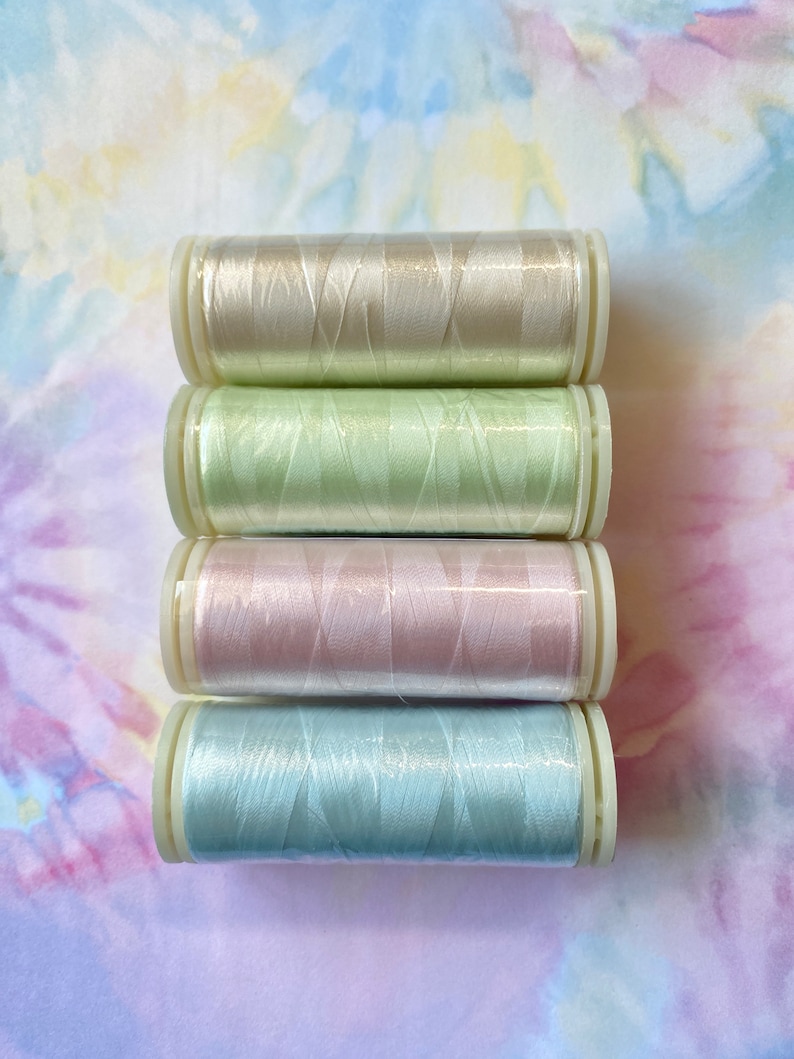 Op de afbeelding: Vier spoelen garen in pastelkleuren: beige, lichtgroen, roze en lichtblauw. De spoelen zijn op elkaar gestapeld.