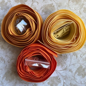 May include: Three spools of fabric trim in shades of orange and yellow. Each spool is coiled and contains a zipper and other hardware in a clear plastic bag. The trim is likely for sewing or crafting projects.
