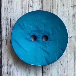 May include: A large, round teal button with two holes. The button has a marbled texture and a glossy finish. The button is set against a light wood grain background.