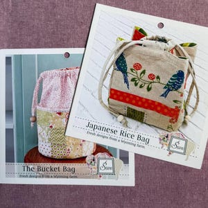 May include: Two instructional pattern cards for fabric bags. One card shows a patchwork bucket bag with the text The Bucket Bag. The other card displays a beige drawstring rice bag with a blue bird and red flower print, labeled Japanese Rice Bag.