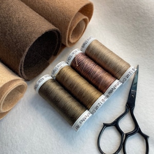 May include: Four spools of brown thread with white labels and a pair of black scissors on a white surface. The labels on the thread spools read "Long Staple 46m/50yd", "Cotton Petites Art. 12 Wt. Cot", "Cotton Petites Art. 12 Wt. Cot", and "Cotton Petites Art. 12 Wt. Cot".