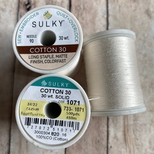 May include: Two spools of white cotton thread, one labeled "Sulky Cotton 30 30 wt. Solid col: 1071" with a barcode and the text "733-1071 500yds 450m 100%CO (Cotton)" and the other labeled "Sulky Cotton 30 Long Staple, Matte Finish, Colorfast 30 wt."