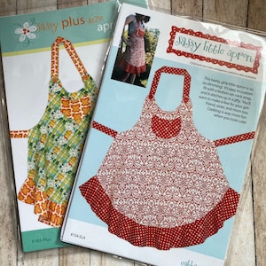 May include: Two sewing patterns for aprons, one for a plus size apron and one for a little apron. The plus size apron pattern is green and orange with a plaid design. The little apron pattern is red with a white floral design. Both patterns have a ruffle at the bottom.