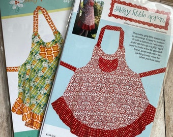 May include: Two sewing patterns for aprons, one for a plus size apron and one for a little apron. The plus size apron pattern is green and orange with a plaid design. The little apron pattern is red with a white floral design. Both patterns have a ruffle at the bottom.