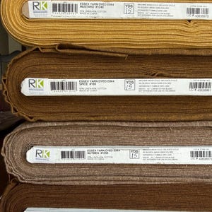 May include: Four stacked bolts of Essex yarn-dyed linen-cotton blend fabric in mustard, spice, nutmeg, and cinnamon colors. Each bolt features a white label with a barcode, product name, and care instructions, noting a 43-inch width for the fabric.