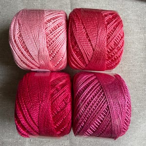 May include: Four balls of embroidery thread arranged in a square grid. The top left ball is light pink, the top right is bright red, the bottom left is deep magenta, and the bottom right is a vibrant purple-pink. Each ball has a smooth texture.