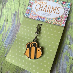 May include: A bee-shaped charm with a silver clasp, featuring a yellow and black design. The charm is attached to a green card with the words "happy CHARMS" and "by Lori Holt of Bee in my Bonnet". The background is a distressed green wood.