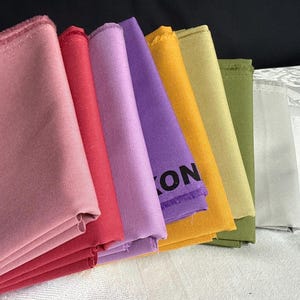 May include: A collection of folded fabric swatches in a spectrum of colors including dusty rose, deep red, light purple, vibrant yellow, muted gold, olive green, and light gray. The purple fabric features partial black text reading ON.