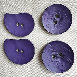 May include: Four purple buttons arranged in a grid. Two buttons are circular with a textured, brushstroke-like surface, and two buttons are asymmetrical, teardrop-shaped with a smooth finish. Each button features two central holes for sewing.
