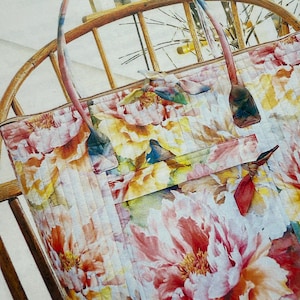 May include: A floral tote bag with a quilted design. The bag has a large, pink peony flower in the center, surrounded by smaller flowers in shades of yellow, orange, and pink. The bag has brown leather handles.