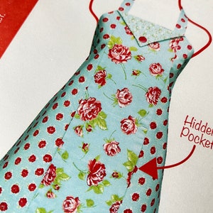 May include: A light blue apron with a red and white floral print and red polka dots. The apron has a hidden pocket on the side. The text "Hidden Pocket" is written on the right side of the image.