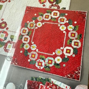 May include: A red and green Christmas quilt with a wreath design. The quilt is made of fabric scraps and has a white background.