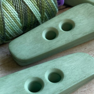 May include: Two sage green plastic buttons with two holes each. The buttons have a wood grain texture.