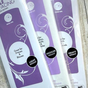 Op de afbeelding: Drie verpakkingen Pellon opstrijkbare vlieseline in lichte, medium en zware gewichten. Elke witte verpakking heeft een paarse branding met tekst waaronder Iron-On, Sewable, Machine Washable en Non-Woven Poly Blend, gemaakt in de VS.