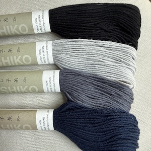 May include: Four skeins of 100% cotton sashiko thread in black, light gray, dark gray, and navy blue. Each skein features a tan paper label printed with the text Sashiko, 20m, Cotton 100%, and Made in Japan, wrapped around the bundled thread.