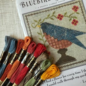 May include: A cross-stitch pattern titled "Bluebird Garden" with a bluebird design, accompanied by a selection of colorful embroidery floss. The floss colors include shades of blue, orange, red, and green, perfect for crafting.