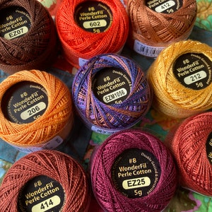 May include: A collection of WonderFil #8 Perle Cotton thread spools in various colors, including brown, orange, purple, and gold. Each spool is labeled with the thread number and weight, 5g. The spools are arranged on a colorful, patterned surface.