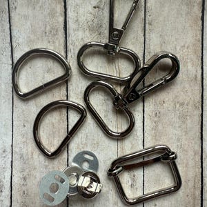 May include: A collection of silver-toned metal hardware components including two D-rings, two swivel lobster claw clasps, one rectangular loop, and a set of circular magnetic snap fasteners with metal backing plates for sewing and craft projects.
