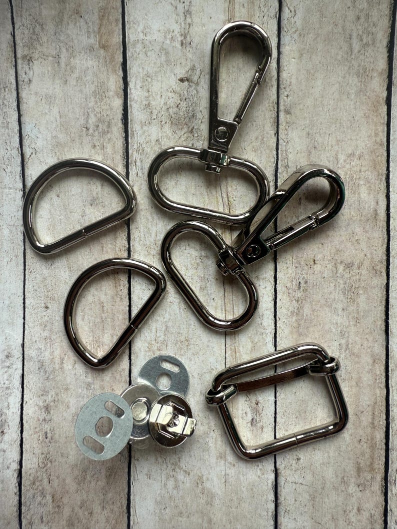 May include: A collection of silver-toned metal hardware components including two D-rings, two swivel lobster claw clasps, one rectangular loop, and a set of circular magnetic snap fasteners with metal backing plates for sewing and craft projects.