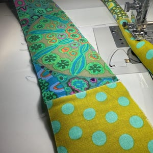 May include: A strip of fabric featuring a vibrant floral pattern in shades of green, blue, and orange, sewn to a mustard yellow fabric with light blue polka dots. The fabric is positioned under the presser foot of a white sewing machine.