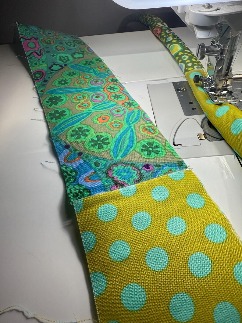 May include: A strip of fabric featuring a vibrant floral pattern in shades of green, blue, and orange, sewn to a mustard yellow fabric with light blue polka dots. The fabric is positioned under the presser foot of a white sewing machine.