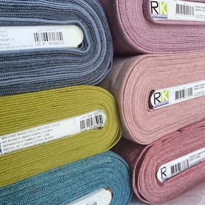K&ouml;nnte beinhalten: F&uuml;nf gestapelte Stoffballen aus garngef&auml;rbtem Robert Kaufman Essex-Stoff in den Farben Blau, Chartreuse, Mauve, Altrosa und Rot. Jede Stoffrolle hat ein wei&szlig;es Etikett mit einem Barcode, Produktinformationen und Pflegehinweisen in Schwarz.