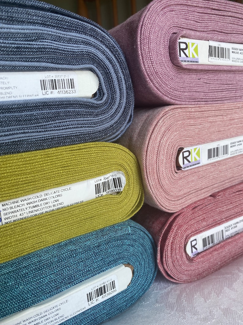 K&ouml;nnte beinhalten: F&uuml;nf gestapelte Stoffballen aus garngef&auml;rbtem Robert Kaufman Essex-Stoff in den Farben Blau, Chartreuse, Mauve, Altrosa und Rot. Jede Stoffrolle hat ein wei&szlig;es Etikett mit einem Barcode, Produktinformationen und Pflegehinweisen in Schwarz.